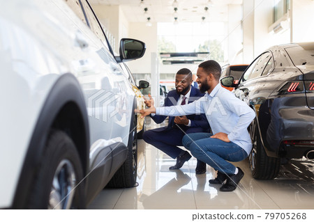 Young Black Man Choosing New Car In Dealership Center, Examining Tires 79705268
