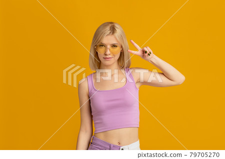 Summer mood. Trendy young lady in glasses making peace sign, isolated over yellow wall, copy space Summer mood. Trendy young lady in glasses making peace sign, isolated over yellow wall, copy space 79705270