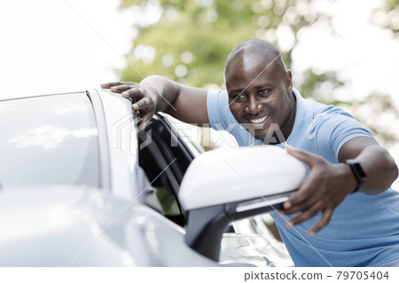 Cheerful black man standing by white car, looking at mirror Cheerful black man standing by white car, looking at mirror 79705404