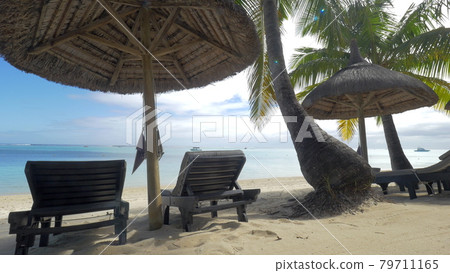 View of empty chaise-longue near native sun umbrella and palm trees against blue water, Mauritius Island View of empty chaise-longue near native sun umbrella and palm trees against blue water, Mauritius Island 79711165