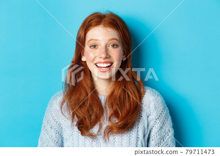Close-up of happy redhead girl in sweater, looking at camera with hopeful smile, standing against blue background 79711473