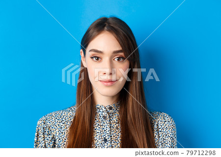 Hopeful candid woman with long hair and makeup, looking at camera with smile, standing against blue background 79712298
