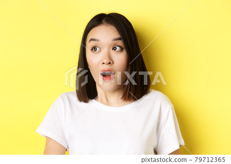 Close up of surprised asian girl drop jaw, gasping and looking left at logo, standing over yellow background Close up of surprised asian girl drop jaw, gasping and looking left at logo, standing over yellow background 79712365