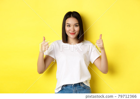 Pretty young asian woman in white t-shirt, showing thumbs up and smiling, praise good offer, recommend product, standing satisfied over yellow background Pretty young asian woman in white t-shirt, showing thumbs up and smiling, praise good offer, recommend product, standing satisfied over yellow background 79712366