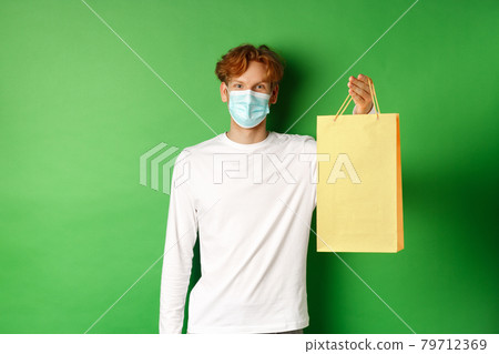 Portrait of handsome boyfriend in face mask giving shopping bag with valentines gift, smiling with eyes, standing over green background 79712369