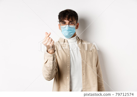 Covid-19, health and real people concept. Cute young man in medical mask showing finger heart and look at camera, standing on white background 79712574