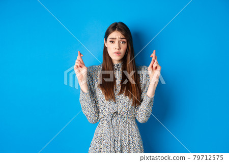 Nervous young woman in dress cross fingers and biting lip worried, praying or making wish, standing on blue background 79712575