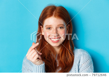 Headshot of cute caucasian woman with red hair and freckles showing heart sign and smiling, standing against blue background 79712646