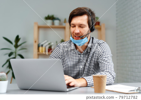 Young man working on laptop with headset and face mask at home 79712782