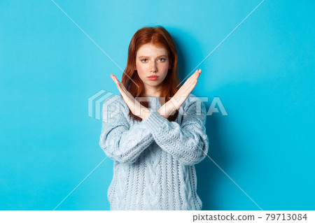 Serious redhead girl looking confident, showing cross gesture to stop and forbid action, standing over blue background Serious redhead girl looking confident, showing cross gesture to stop and forbid action, standing over blue background 79713084