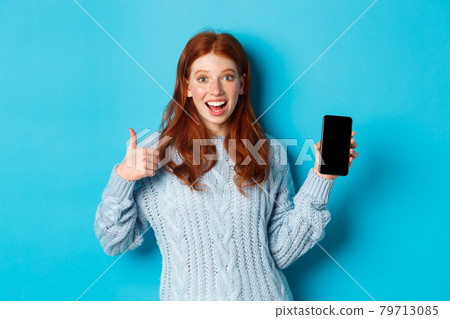 Smiling redhead girl showing smartphone screen, holding phone and demonstrating app, making thumb up in approval, recommending online store, blue background Smiling redhead girl showing smartphone screen, holding phone and demonstrating app, making thumb up in approval, recommending online store, blue background 79713085