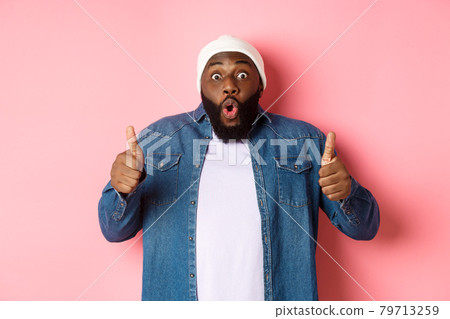 Impressed african-american hipster guy showing thumbs-up, approve and like offer, praising great choice, standing over pink background 79713259