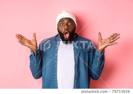 Shocked and impressed Black man staring at camera with complete disbelief, saying wow, standing in beanie and hipster shirt over pink background 79713656