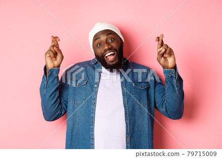 Optimistic african-american man making wish, holding fingers crossed and smiling, standing over pink background Optimistic african-american man making wish, holding fingers crossed and smiling, standing over pink background 79715009