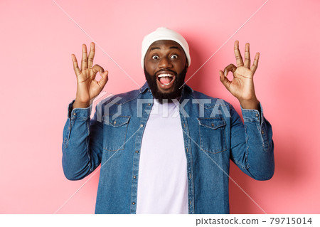 Impressed Black man in beanie and shirt, showing ok signs and staring at camera amazed, smiling happy, praise great offer, recommending product, pink background Impressed Black man in beanie and shirt, showing ok signs and staring at camera amazed, smiling happy, praise great offer, recommending product, pink background 79715014