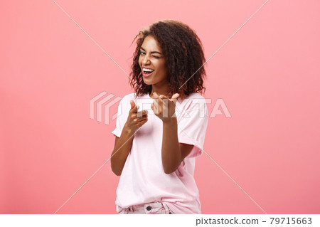 Hey you awesome. Playful charming and happy carefree dark-skinned trendy girl with curly hairstyle winking and smiling broadly making finger gun move towards camera checking out cool outfit Hey you awesome. Playful charming and happy carefree dark-skinned trendy girl with curly hairstyle winking and smiling broadly making finger gun move towards camera checking out cool outfit 79715663