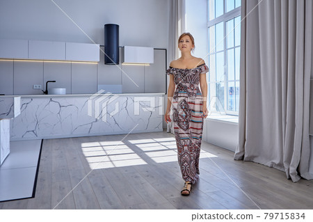 Caucasian woman over 40 walks through studio apartment in long summer dress. 79715834