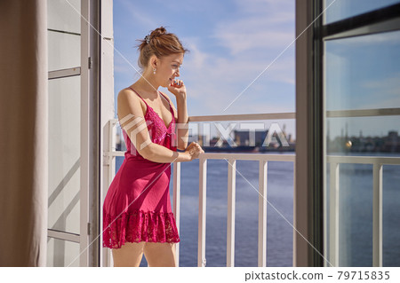 Woman in peignoir stands on loggia and looks out into street. Woman in peignoir stands on loggia and looks out into street. 79715835