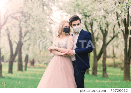 Young loving couple walking in medical masks in the park during quarantine on their wedding day. Young loving couple walking in medical masks in the park during quarantine on their wedding day. 79720012