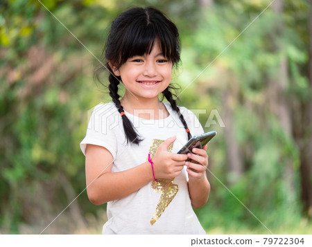 A seven-year-old Asian girl is smiling happily as she uses her mobile phone to search for information while on a family trip to the park. 79722304