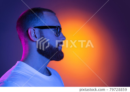 Close up portrait of young caucasian bearded man in sunglasses isolated on dark studio background in neon lights. Side view Close up portrait of young caucasian bearded man in sunglasses isolated on dark studio background in neon lights. Side view 79728859