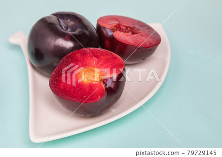 On a white saucer is a large plum, cut in half, and next to it is a whole plum. On a white saucer is a large plum, cut in half, and next to it is a whole plum. 79729145