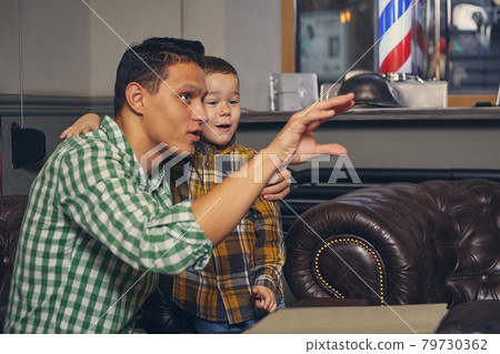Young father and his stylish little son in the barbershop in the waiting room. Young father and his stylish little son in the barbershop in the waiting room. 79730362
