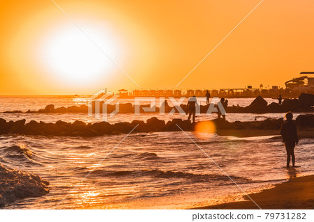 Orange sunset sky with a skyline on the sea coast with stones breakwater and holidaymakers in the resort area 79731282