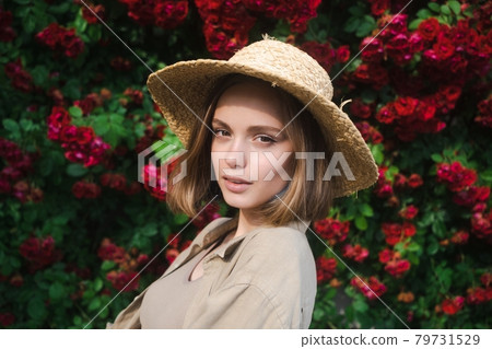 Elegant beautiful blonde young woman in a straw hat on the background of a bush of blooming roses. 79731529