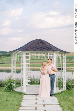 Happy mature couple in elegant clothes, standing near wooden gazebo on the shore of beautiful lake Happy mature couple in elegant clothes, standing near wooden gazebo on the shore of beautiful lake 79738365