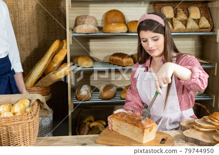 The saleswoman starts cutting the bread and the baker arranges the rolls and other goods in the store. Family bakery and shop. 79744950