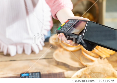 The shop assistant accepts payment via the mobile phone for the sold bread. Cashless payment in the bakery store. 79744976