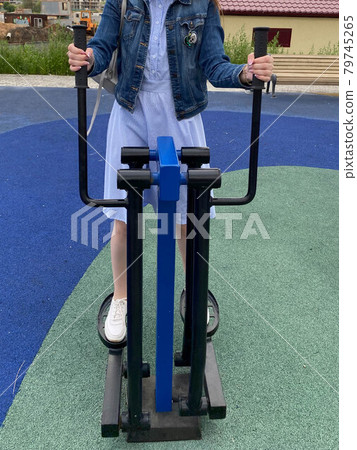 A woman goes in for sports on a new modern blue fitness leg trainer for walking and running on an outdoor workout site A woman goes in for sports on a new modern blue fitness leg trainer for walking and running on an outdoor workout site 79745265