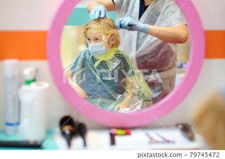 Preschooler boy wearing facemask getting haircut in salon during coronavirus epidemic. Hairdresser examined and treating baby hair for lice. Preschooler boy wearing facemask getting haircut in salon during coronavirus epidemic. Hairdresser examined and treating baby hair for lice. 79745472