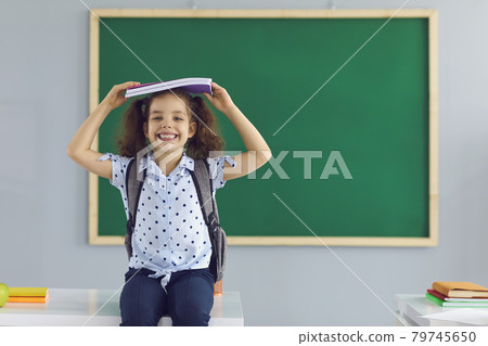 Concept back to school. Happy schoolgirl holds a book over her head laughing standing on the background of a green school board in the classroom. Concept back to school. Happy schoolgirl holds a book over her head laughing standing on the background of a green school board in the classroom. 79745650