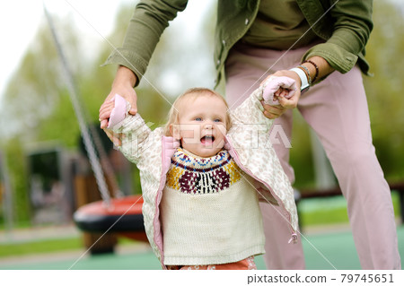 Cute toddler girl having fun on outdoor playground. Young father is teaching his little daughter to walk. First step of baby. Walking for family with little kids. Cute toddler girl having fun on outdoor playground. Young father is teaching his little daughter to walk. First step of baby. Walking for family with little kids. 79745651
