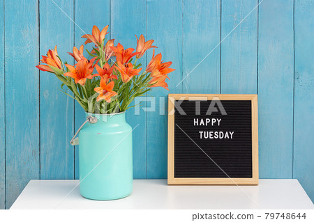 Happy Tuesday words on black letter board and bouquet of orange flowers on table against blue wooden wall. Concept Hello Tuesday Happy Tuesday words on black letter board and bouquet of orange flowers on table against blue wooden wall. Concept Hello Tuesday 79748644