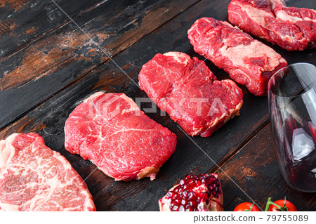 Different raw beef steaks with seasonings and red wine in bottle and glass on old wooden dark planks side view with space for text. Different raw beef steaks with seasonings and red wine in bottle and glass on old wooden dark planks side view with space for text. 79755089