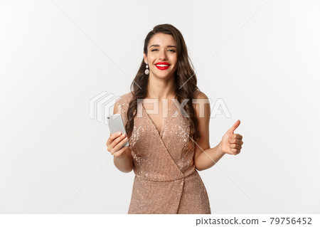 Holidays, online shopping concept. Attractive woman in elegant dress and jewelry, showing thumb up in approval while using mobile phone, smiling satisfied, white background 79756452