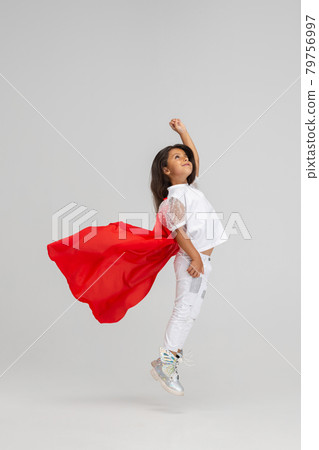 Small superhero. Little cute Caucasian girl in a red cape isolated over white studio background. Side view Small superhero. Little cute Caucasian girl in a red cape isolated over white studio background. Side view 79756997