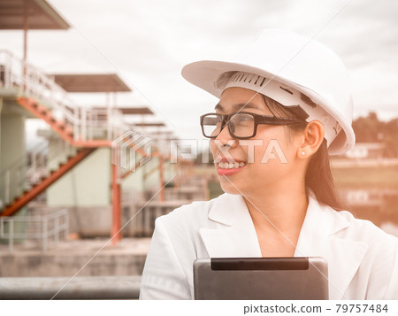 Female engineer in a white helmet talking radio communication (walkie talkie) and holding a digital tablet, stands in construction site of the hydroelectric dam. Female engineer in a white helmet talking radio communication (walkie talkie) and holding a digital tablet, stands in construction site of the hydroelectric dam. 79757484