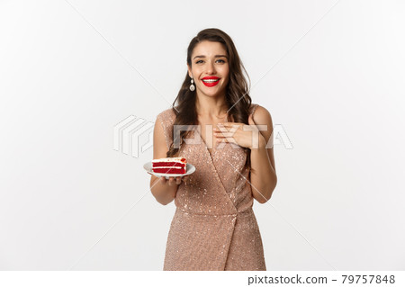 Party and celebration concept. Attractive slim woman in elegant dress holding piece of cake and smiling, standing over white background 79757848