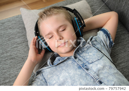 Teen boy in headphones listen to music on couch at home. Teenager enjoy music - blond boy while lying on sofa. Teen boy in headphones listen to music on couch at home. Teenager enjoy music - blond boy while lying on sofa. 79757942