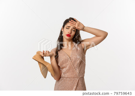 Party and celebration concept. Tired woman in elegant dress take off pair of heels, holding hand on forehead, express fatigue, standing over white background 79759643