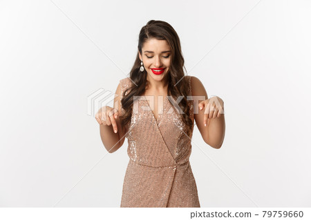 Beautiful young woman with red lipstick, wearing elegant dress for christmas party, pointing fingers down and smiling pleased, standing over white background 79759660