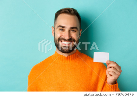 Close-up of handsome caucasian man going on shopping, showing credit card and smiling, standing over turquoise background 79760479