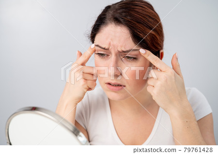 Caucasian woman looks at herself in the mirror and is upset because of facial wrinkles on her face. Caucasian woman looks at herself in the mirror and is upset because of facial wrinkles on her face. 79761428