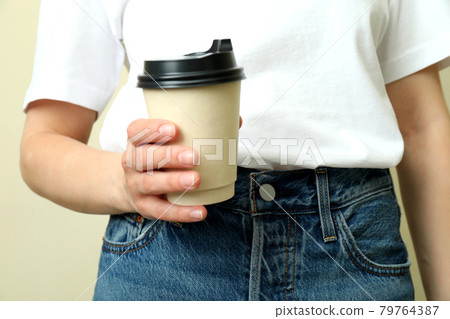 Young woman in t-shirt hold paper cup against beige background 79764387