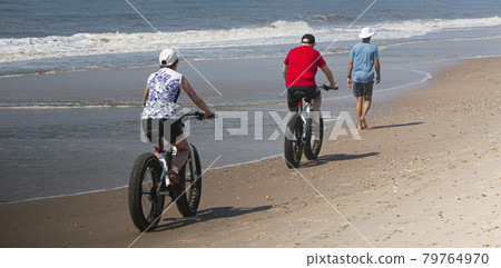 Couple riding fat wheel bikes passing a walker on the waters edge Couple riding fat wheel bikes passing a walker on the waters edge 79764970
