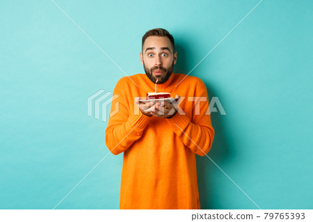 Handsome adult man celebrating birthday, blowing out candle on cake and making wish, standing against turquoise background 79765393